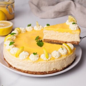 A creamy lemon cheesecake with a graham cracker crust, decorated with piped whipped cream, lemon slices, and mint leaves. One slice is being lifted from the whole cake on a white plate.