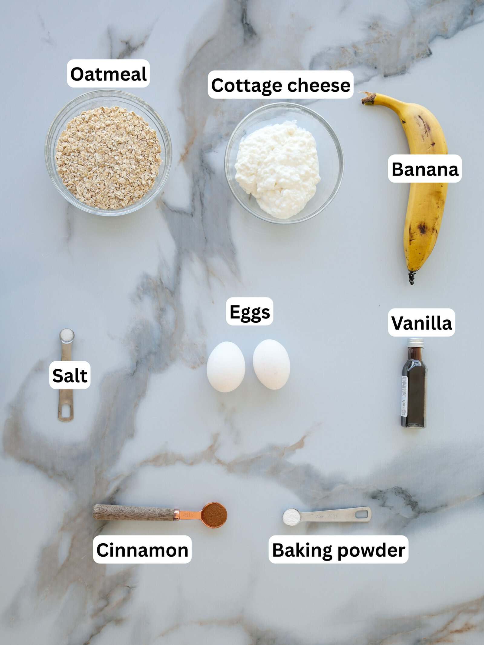 A marble surface displays labeled ingredients: a bowl of oatmeal, a bowl of cottage cheese, a banana, two eggs, a bottle of vanilla, small amounts of salt, cinnamon, and baking powder in measuring spoons.
