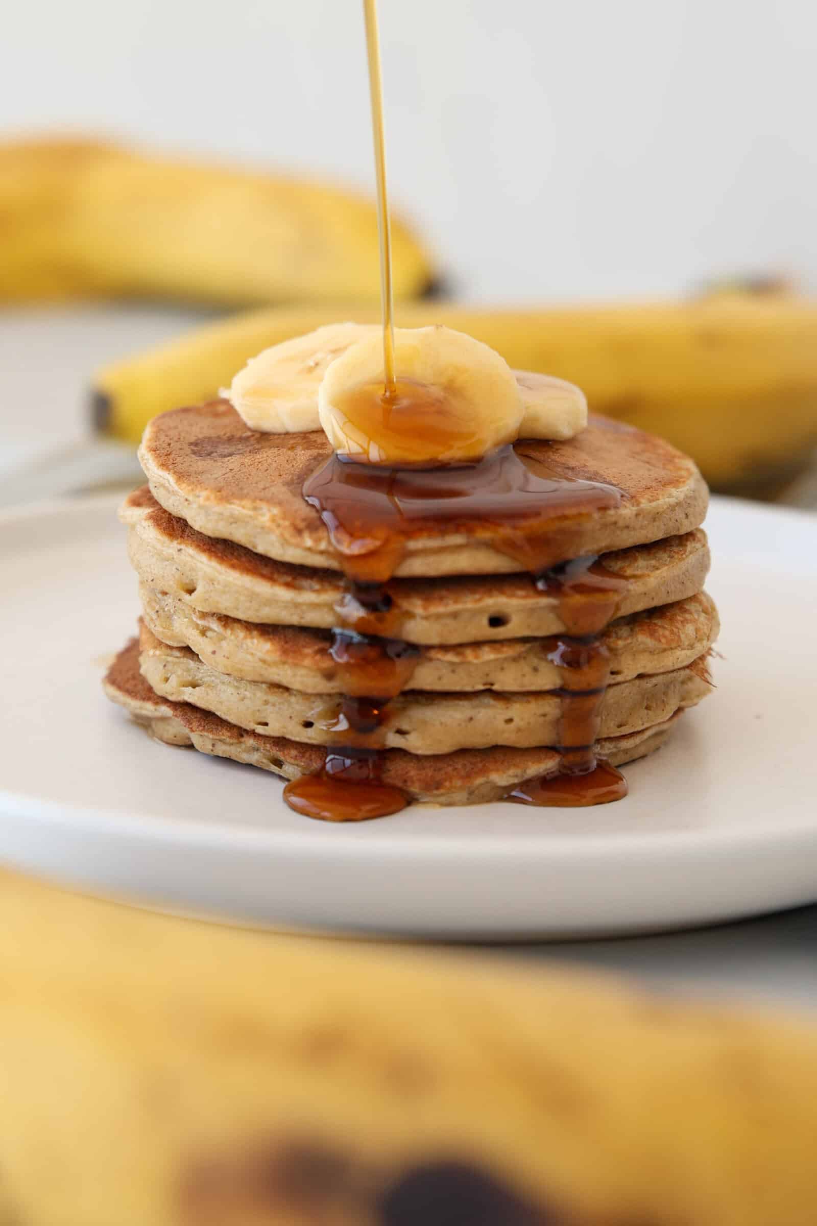 A stack of banana cottage cheese pancakes topped with banana slices and syrup, with more syrup being poured on top. Bananas are visible in the background, and the pancakes are served on a white plate.