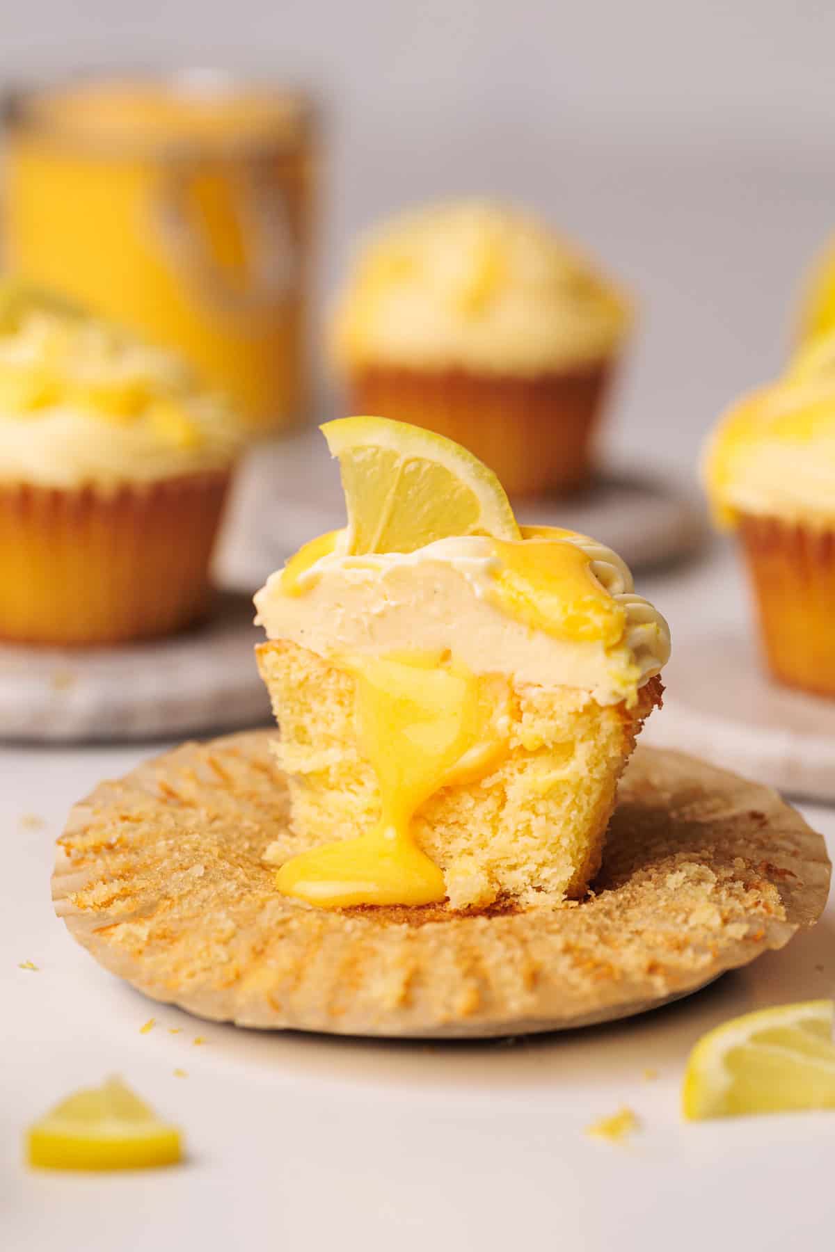 A moist lemon cupcake cut in half reveals a gooey lemon filling inside, topped with creamy frosting and a lemon wedge. Other frosted cupcakes and lemon slices are visible in the background.