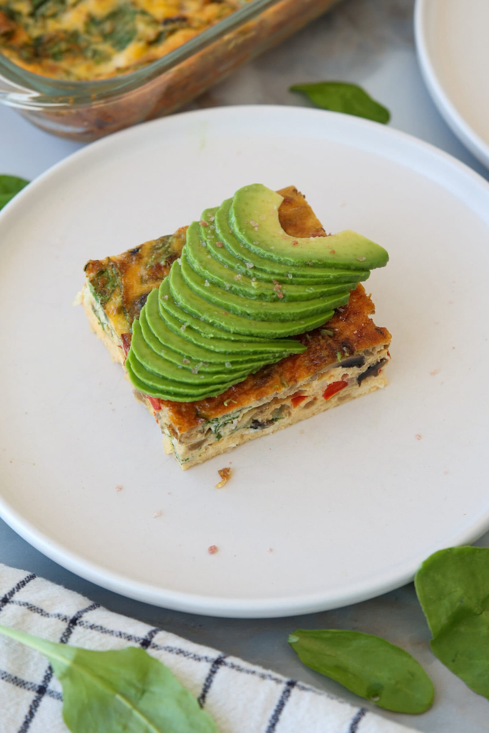 A square slice of high-protein cottage cheese egg casserole topped with fanned avocado slices sits on a white plate, with a few spinach leaves and a striped cloth nearby.