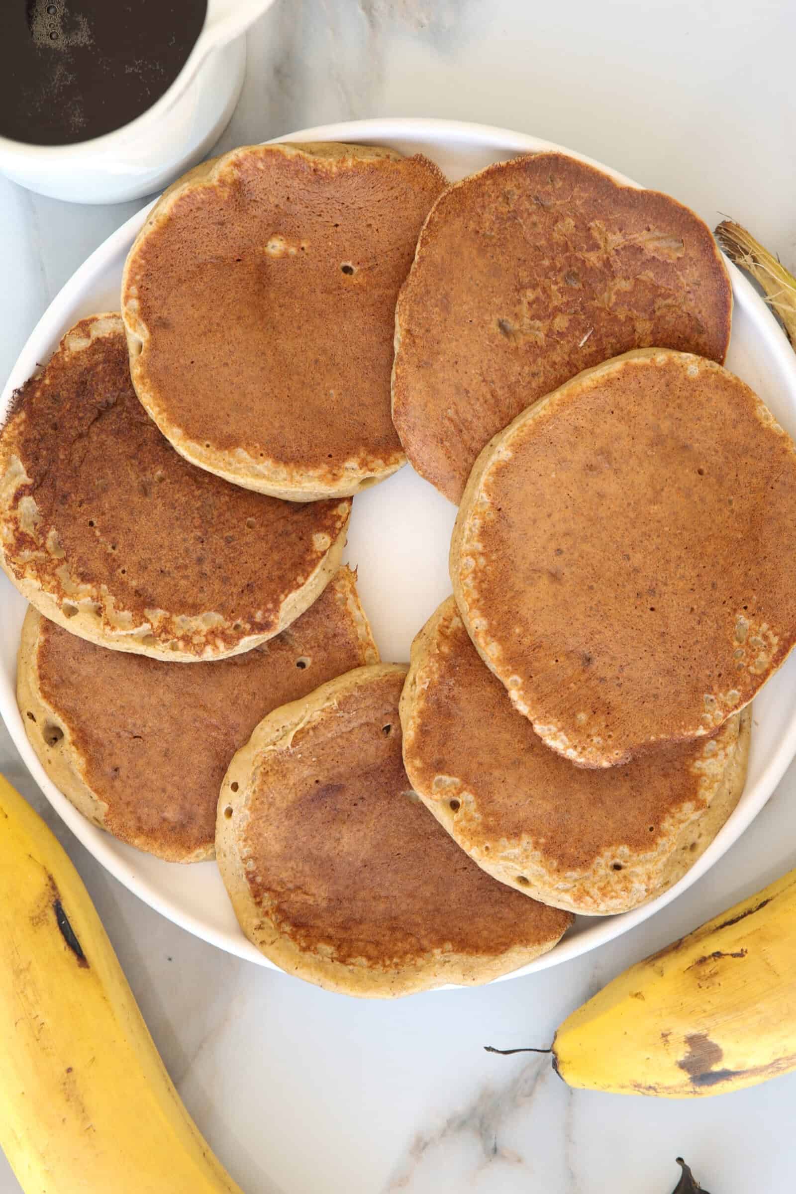 A white plate with seven golden-brown pancakes arranged in a circle, surrounded by ripe bananas and a small container of syrup on a light marble surface.