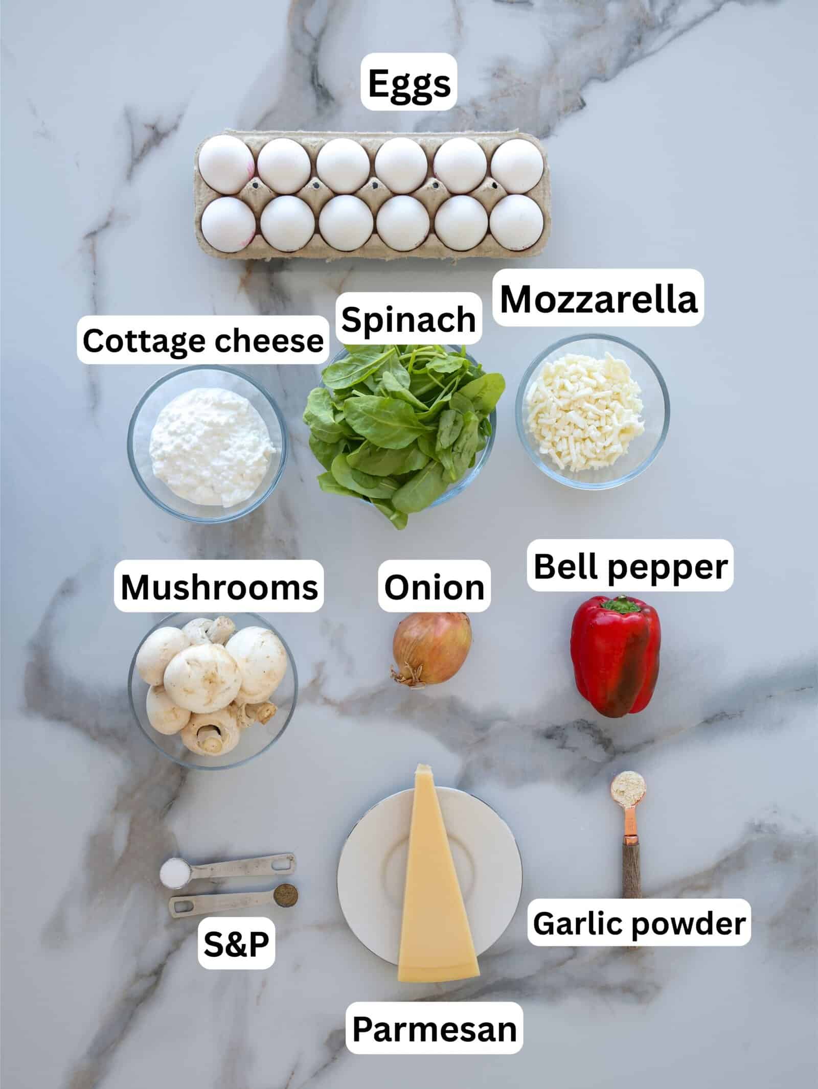 A flat lay of labeled ingredients on a marble surface, including eggs, cottage cheese, spinach, mozzarella, mushrooms, onion, bell pepper, salt and pepper, garlic powder, and a wedge of parmesan cheese.