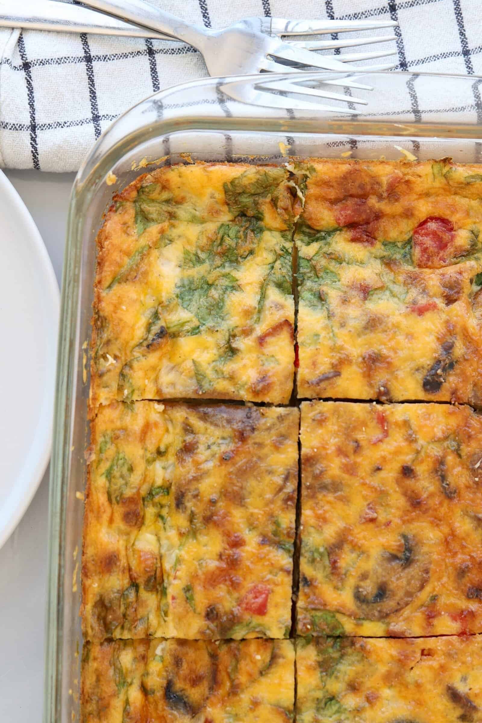 A glass baking dish filled with a baked cottage cheese egg casserole cut into six squares, with visible spinach, tomatoes, and mushrooms. Three forks rest on a white and blue checkered towel in the background.