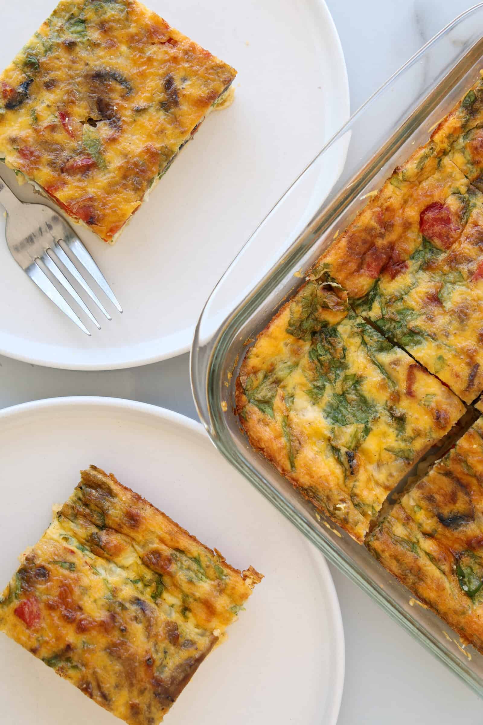A glass baking dish with a colorful baked cottage cheese egg bake cut into squares, with two servings on white plates and a fork next to one piece. The frittata contains visible vegetables and herbs.