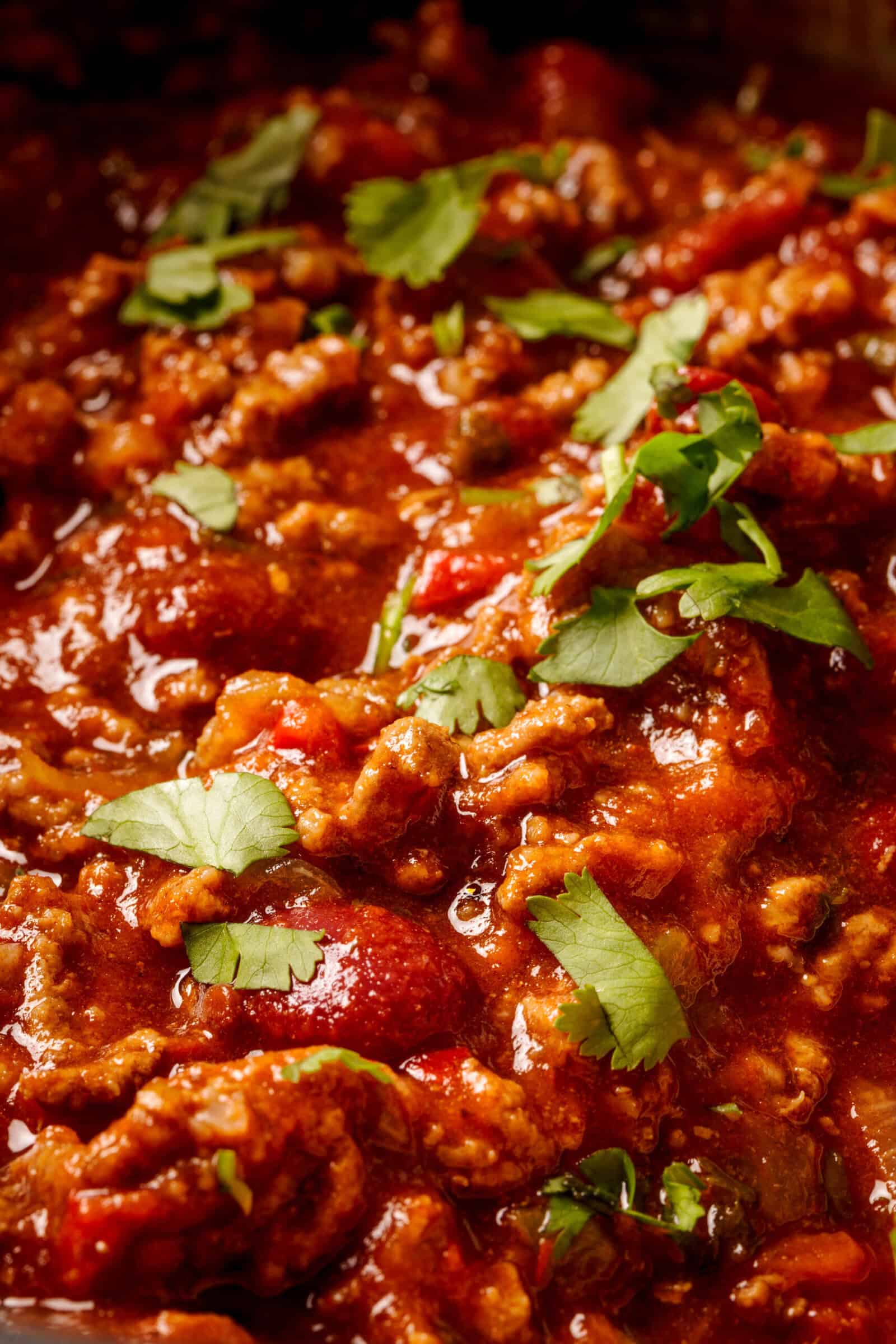 Close-up of a hearty chili dish with ground meat, tomatoes, and beans in a rich red sauce, topped with fresh cilantro leaves.