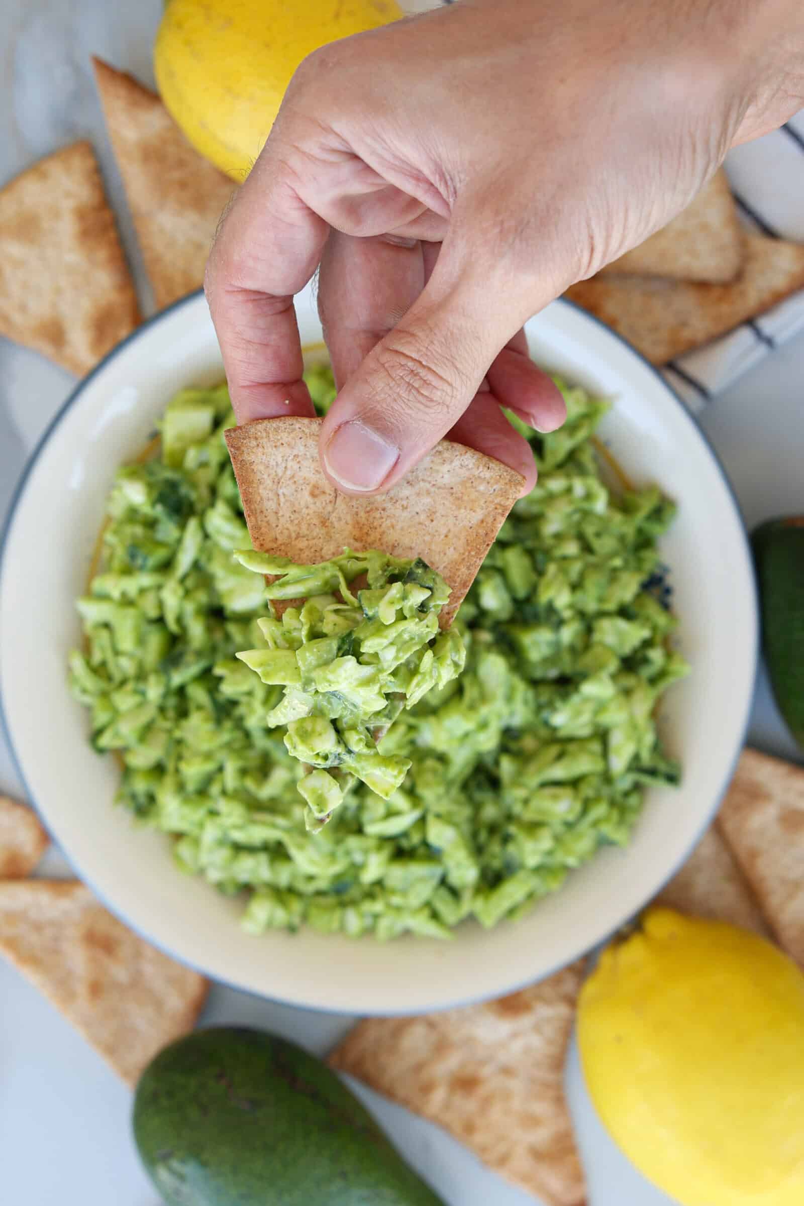 A hand dips a pita chip into a bowl of chunky green goddess salad, surrounded by pita chips, a lemon, and an avocado on a white surface.