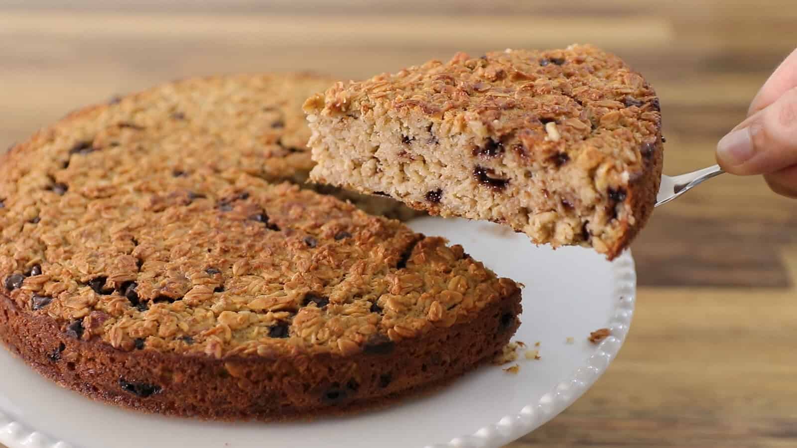 A hand lifts a slice of baked banana oatmeal cake from a round cake on a white plate, showing the cake's dense, moist texture. The background is a wooden surface.