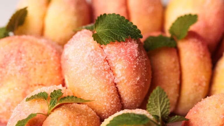 Close-up of peach-shaped cookies covered in sugar, decorated with green mint leaves, arranged closely together.