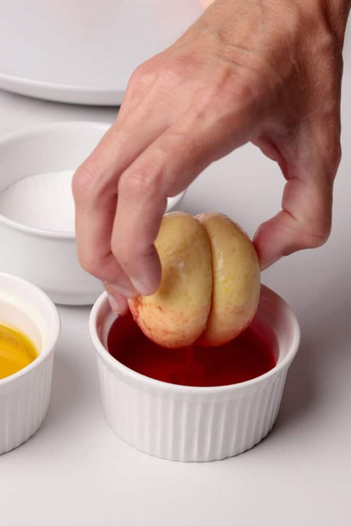A hand dips a round, sandwiched cookie into a small white ramekin filled with red liquid. Nearby are additional ramekins containing yellow liquid and white granulated sugar.