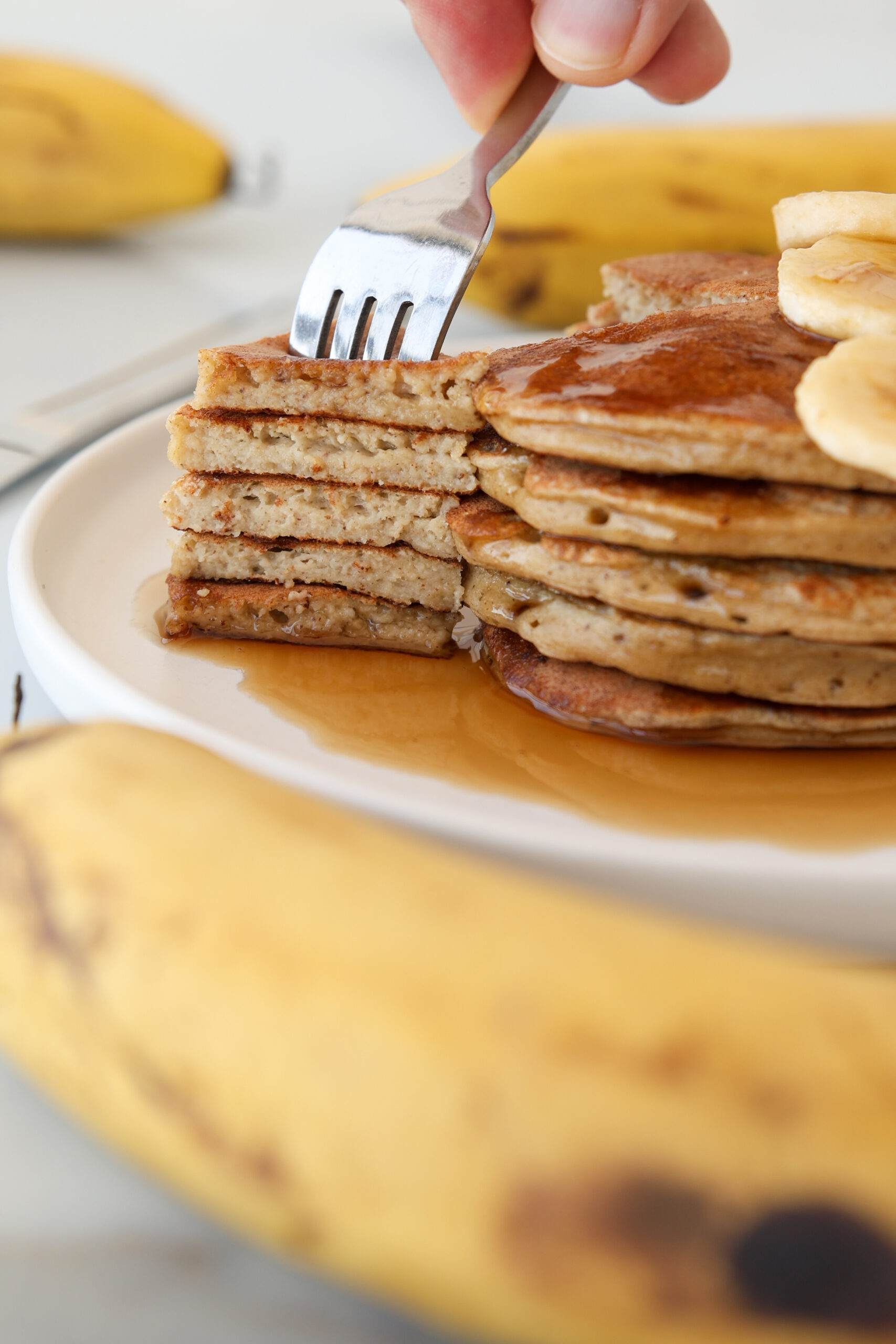 A close-up of a stack of high-protein cottage cheese banana pancakes topped with banana slices and syrup. A hand is using a fork to lift a cut piece. Fresh bananas are visible in the foreground and background.