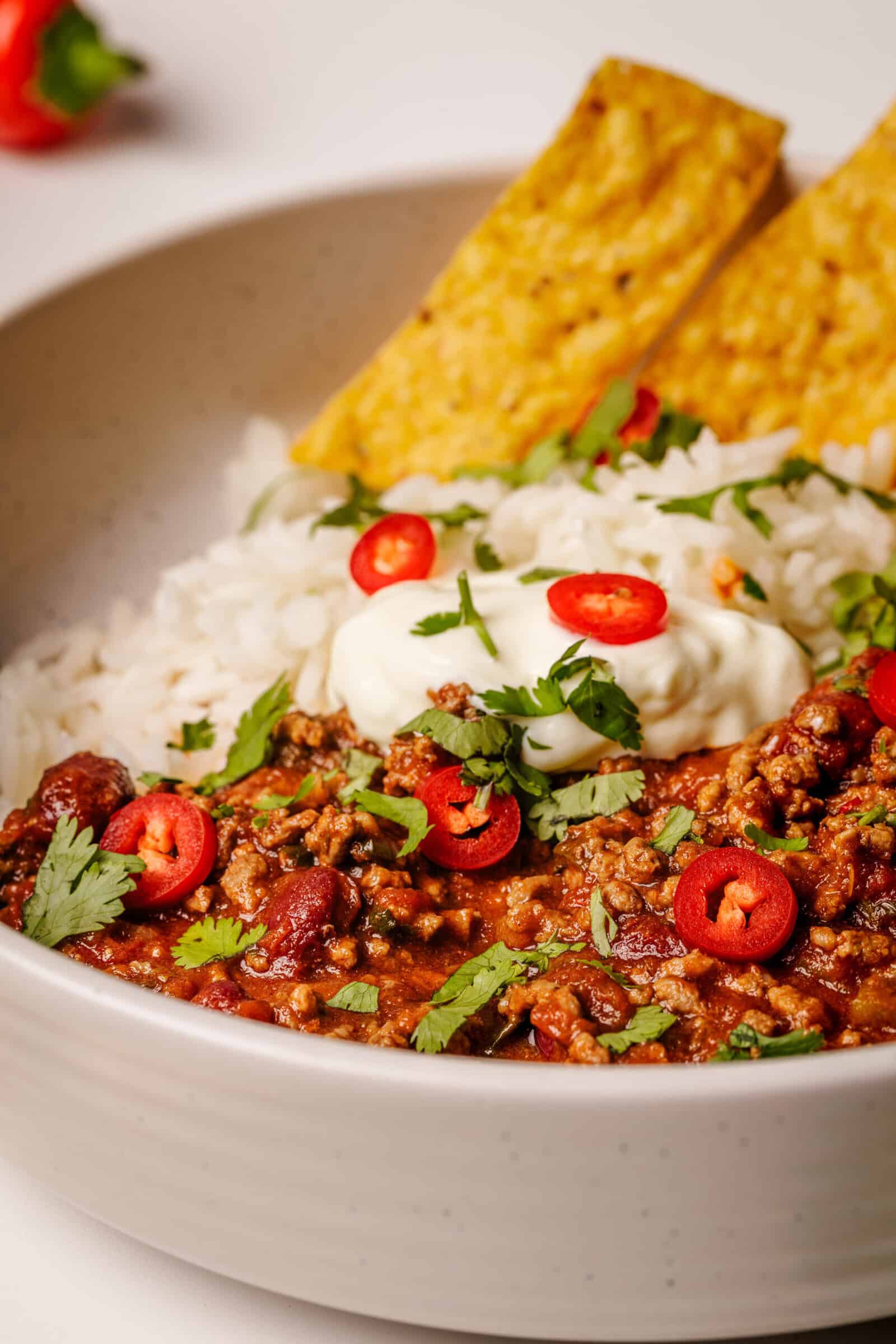 A bowl with white rice, chili con carne topped with sliced red chili and cilantro, a dollop of sour cream, and two yellow tortilla chips.