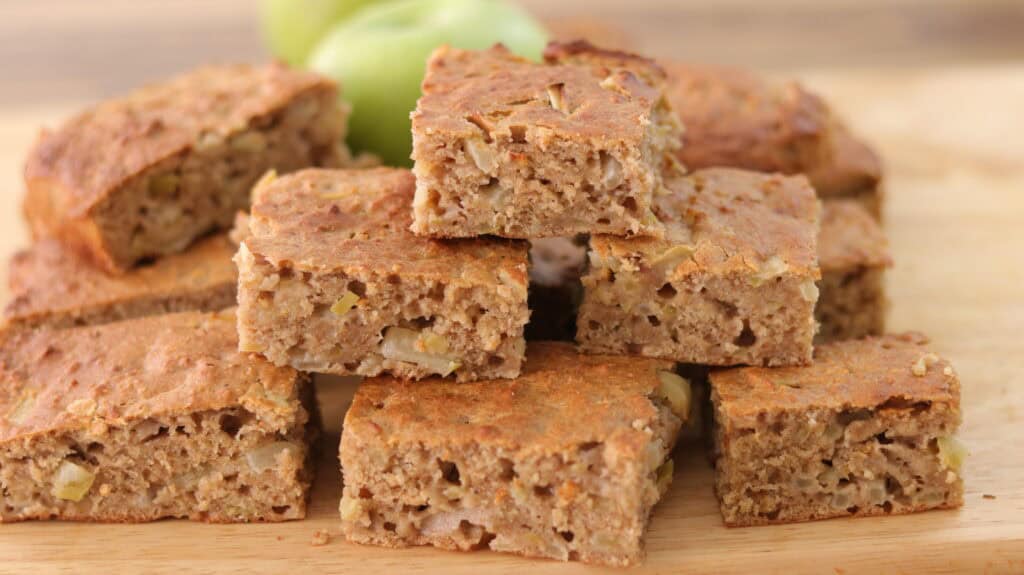 A close-up of several pieces of apple cake stacked on a wooden surface, showing a moist, spongy texture with visible apple chunks. Green apples are blurred in the background.