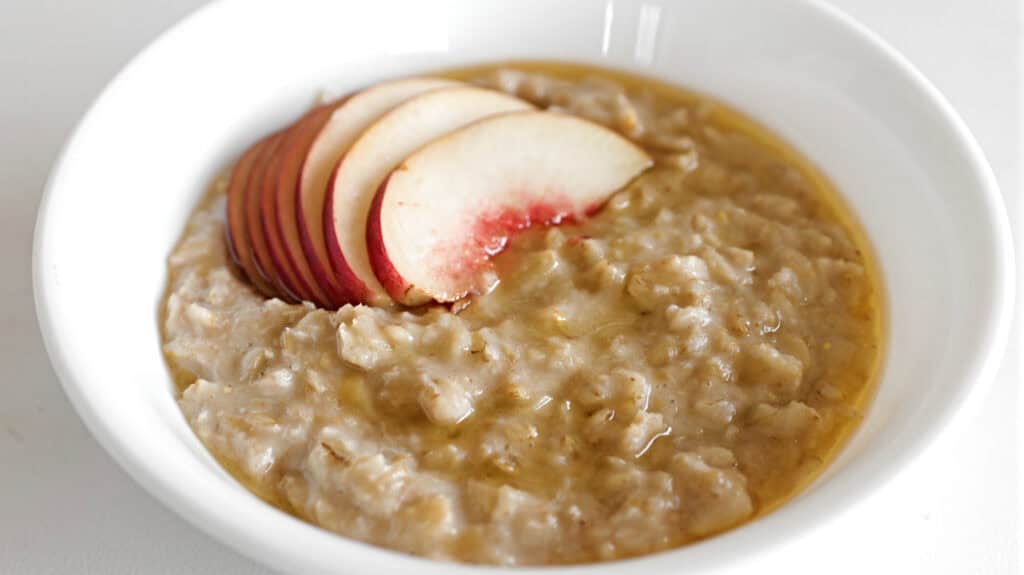 A white bowl filled with oatmeal topped with thinly sliced red apple and drizzled with honey or syrup.