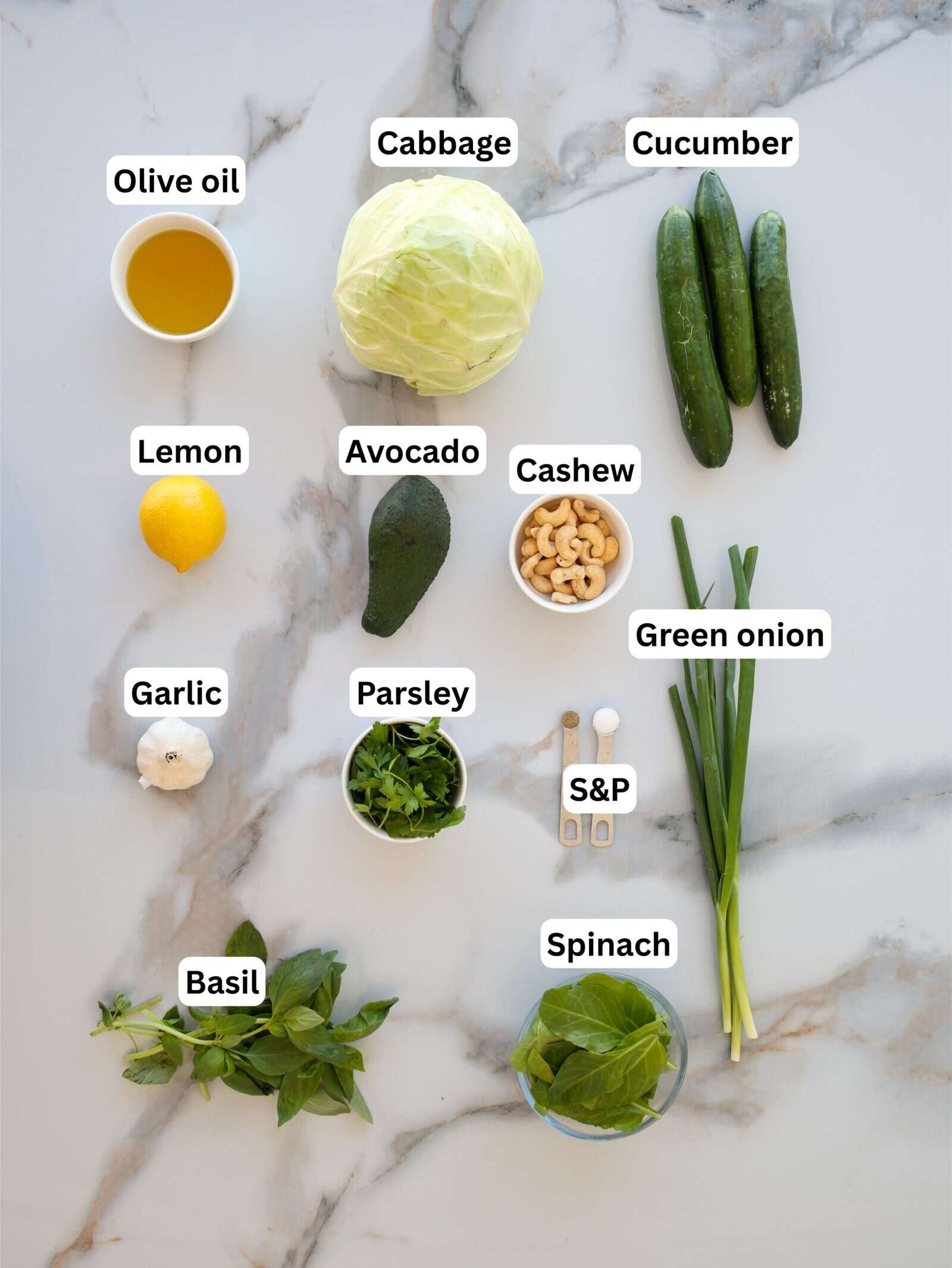 A marble surface displays labeled ingredients: olive oil, lemon, garlic, basil, avocado, parsley, cabbage, cashew nuts, cucumber, green onion, spinach, and salt & pepper in small bowls.