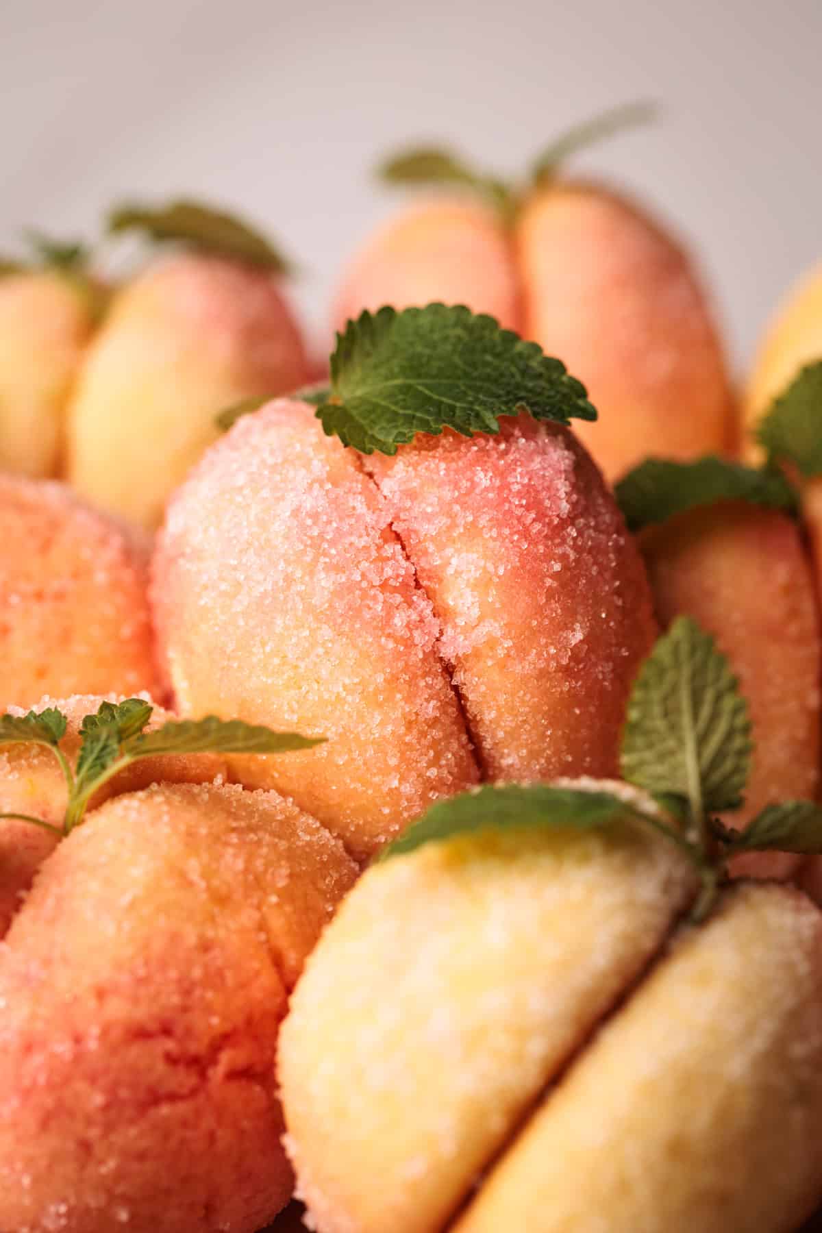 Close-up of sugar-coated Italian peach cookies (Pesche Dolci) with a pink and yellow gradient, decorated with green mint leaves, arranged in a cluster.