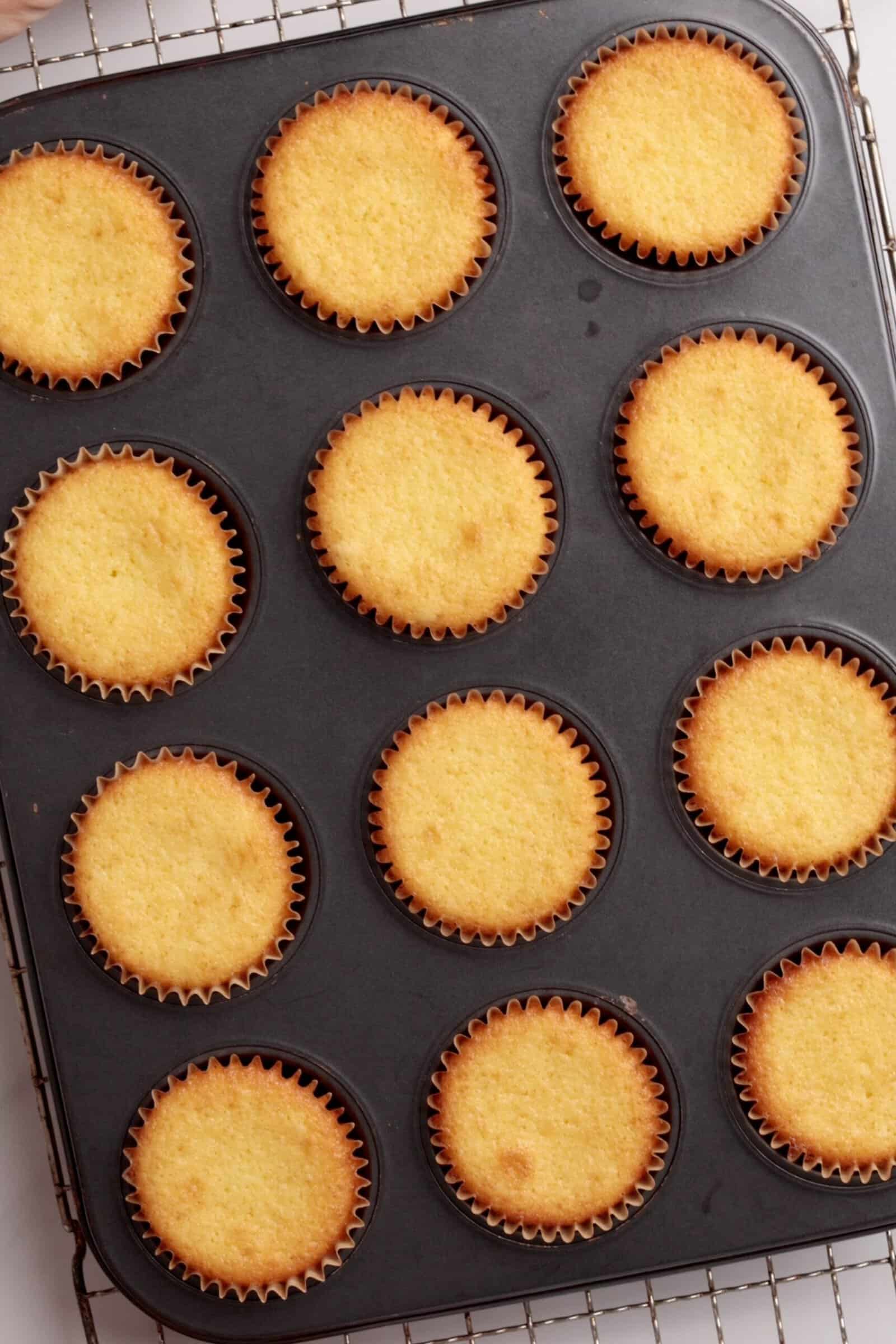 A muffin tin holding twelve golden lemon cupcakes in paper liners, resting on a cooling rack. The cupcakes appear freshly baked, with lightly browned edges and smooth, slightly domed tops.