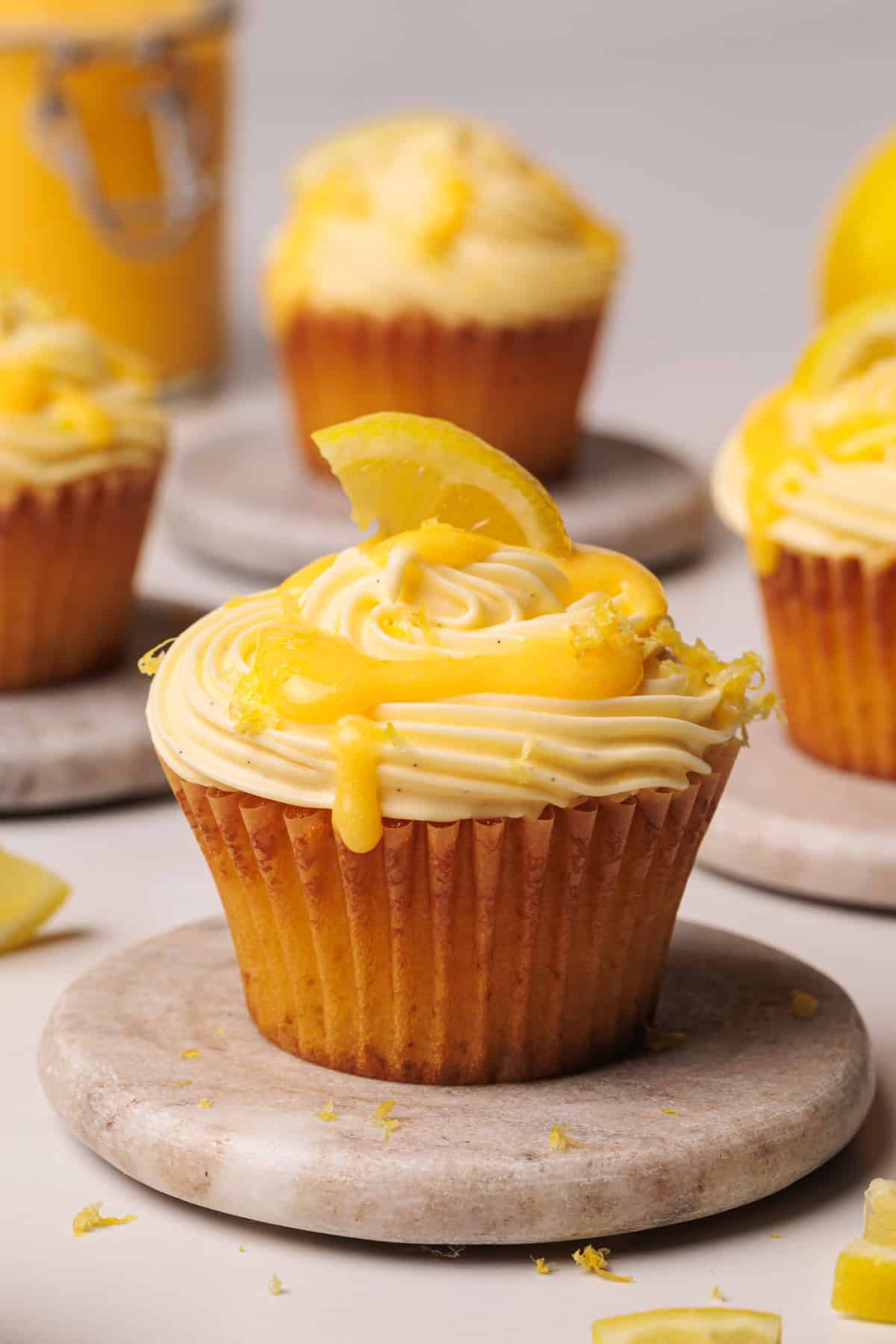 A lemon cupcake with swirled yellow frosting, drizzled with lemon curd, and topped with a lemon slice sits on a marble coaster. More cupcakes are blurred in the background.