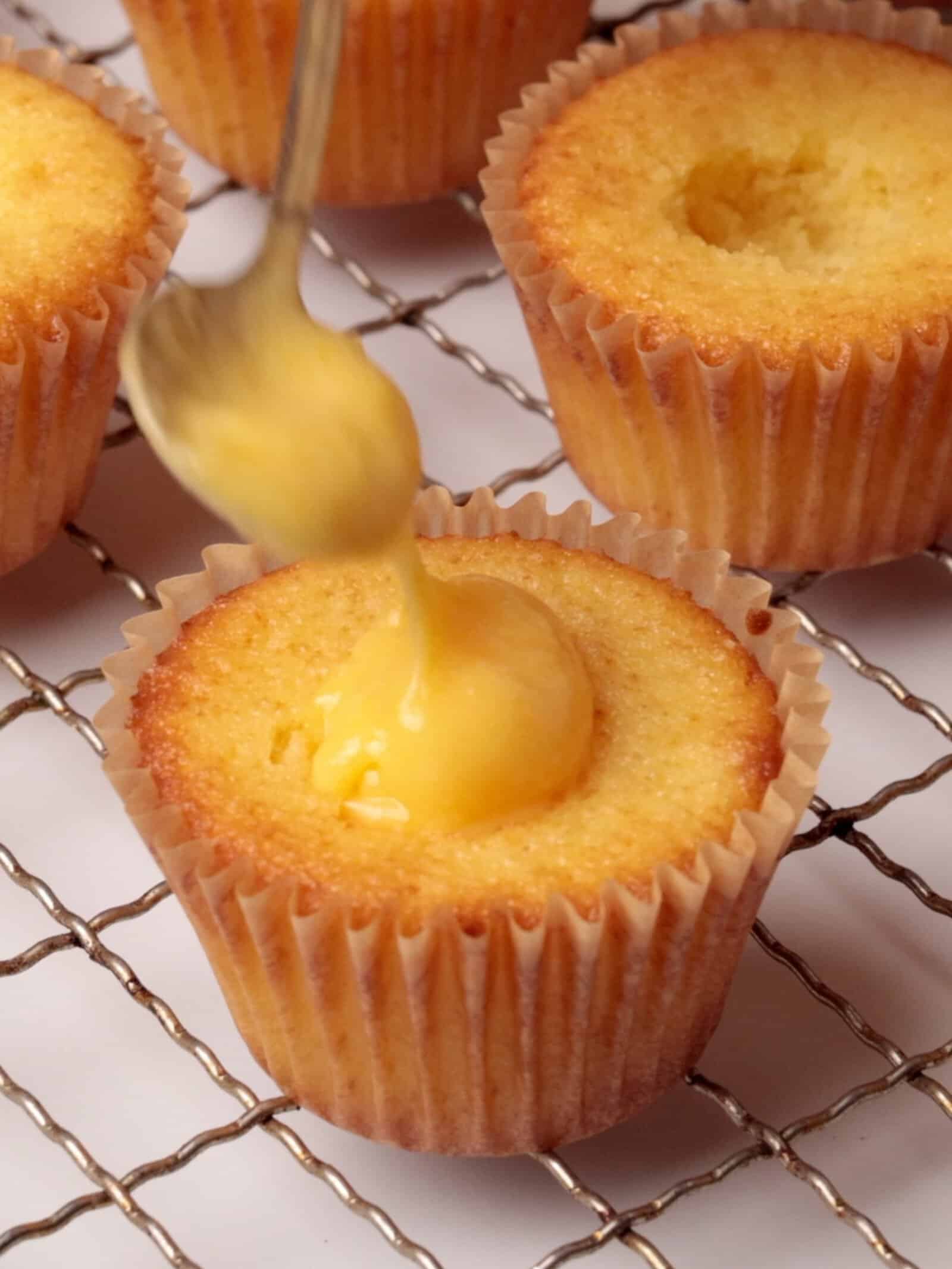 A spoon is filling a hole in a cupcake with yellow lemon curd, while other plain cupcakes cool on a wire rack in the background.