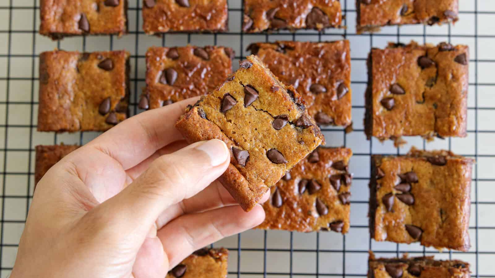 A hand holds a Peanut Butter Banana Chocolate Chip Bar above a cooling rack with more blondie squares arranged neatly in the background.