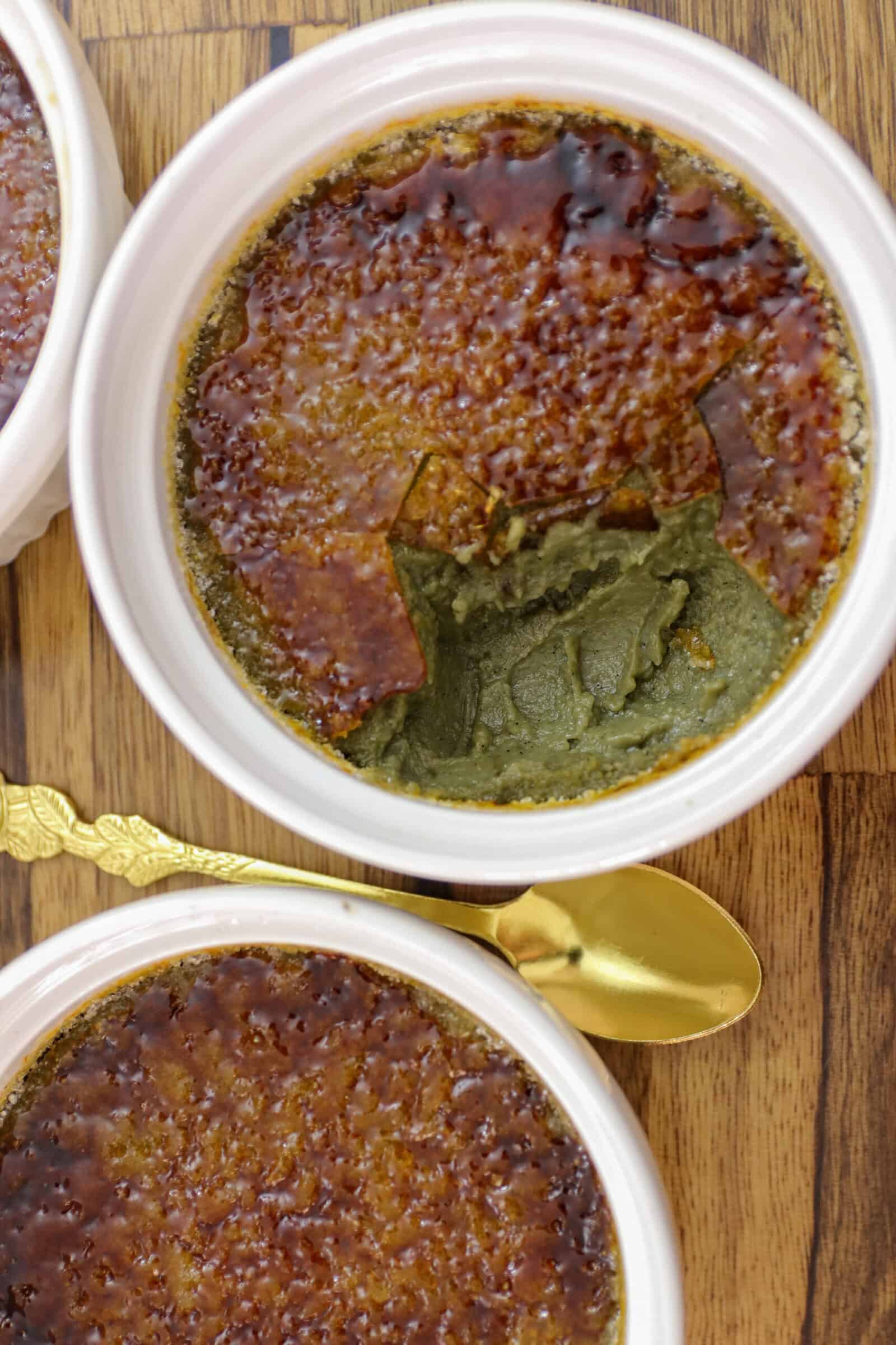 Two ramekins of pistachio creme brulee with caramelized sugar tops, one partially eaten to reveal a green custard inside, placed on a wooden surface with a gold spoon.