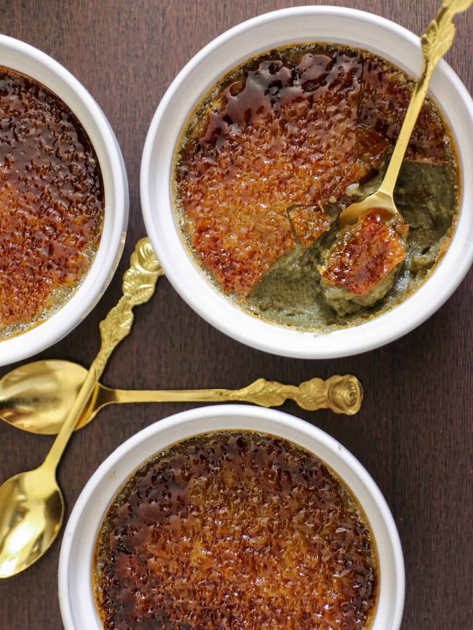 Three ramekins of crème brûlée with caramelized sugar tops sit on a dark surface. One has a spoon breaking through the crust, revealing a creamy interior. Two ornate gold spoons are placed beside the ramekins.