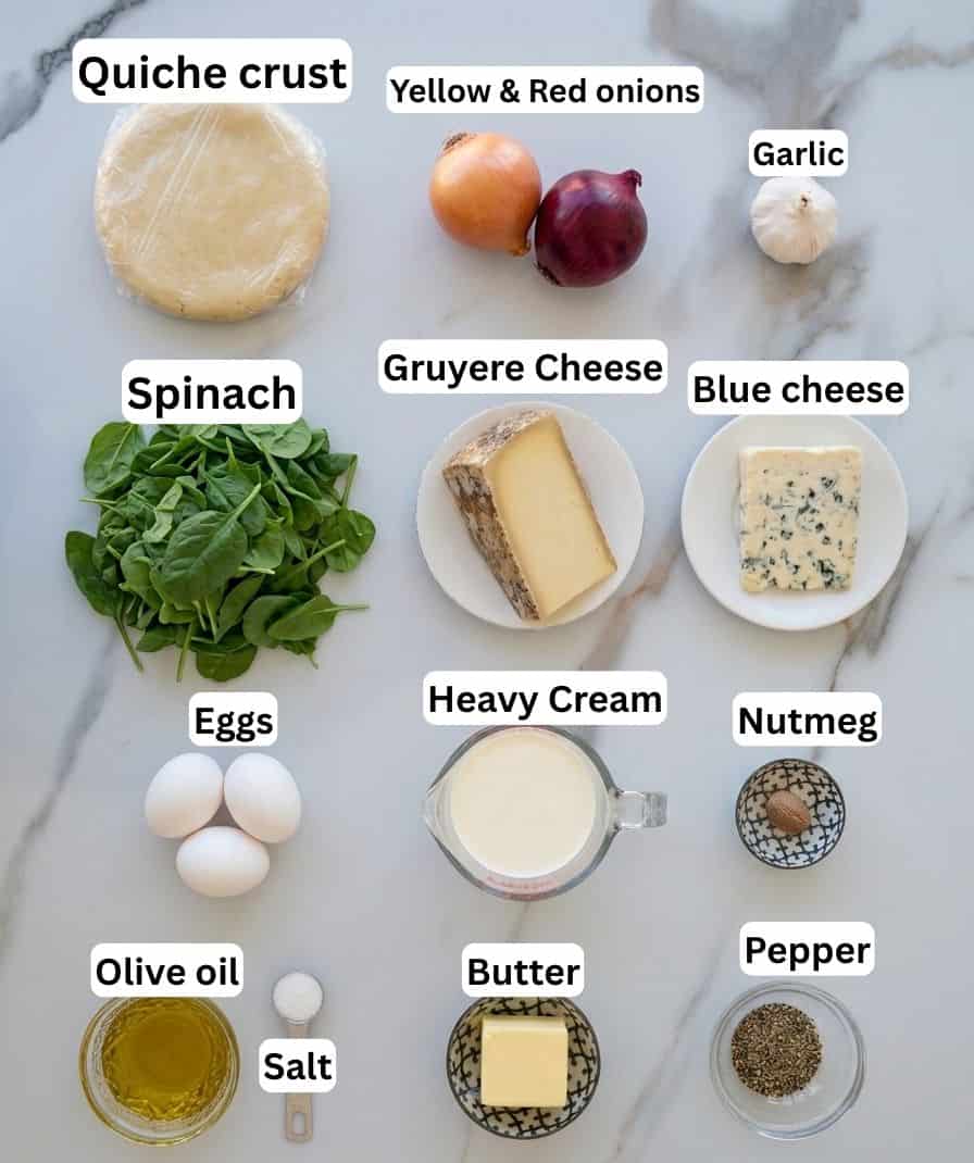 Top-down view of quiche ingredients: quiche crust, yellow and red onions, garlic, spinach, Gruyère cheese, blue cheese, eggs, heavy cream, nutmeg, pepper, olive oil, salt, and butter, all labeled on a marble surface.