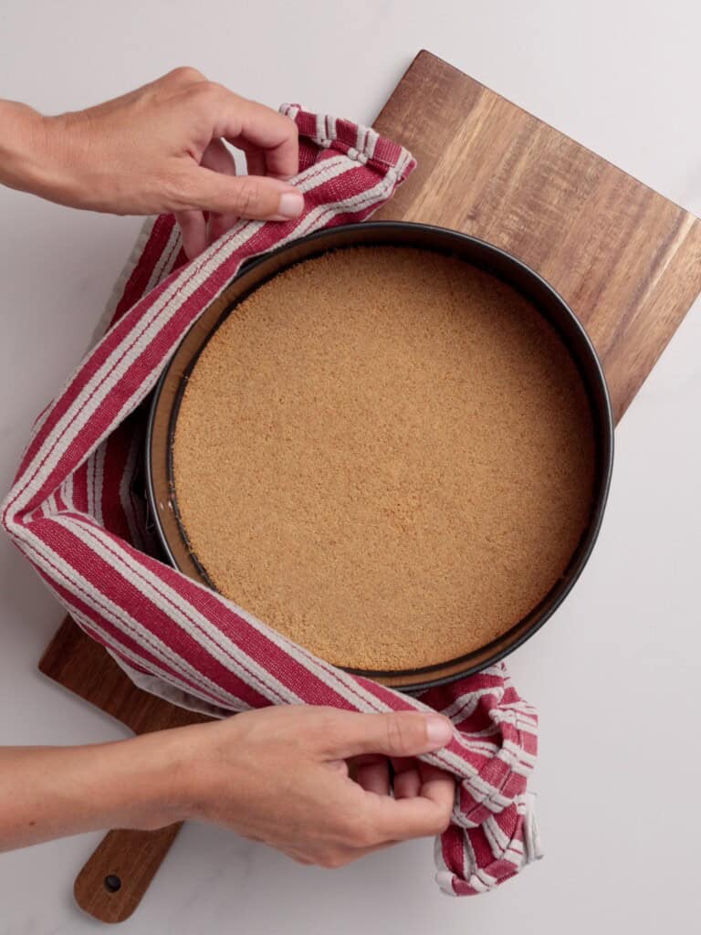 A person wraps a round cake pan filled with a crumbly graham cracker crust in a red and white striped cloth, resting on a wooden cutting board.