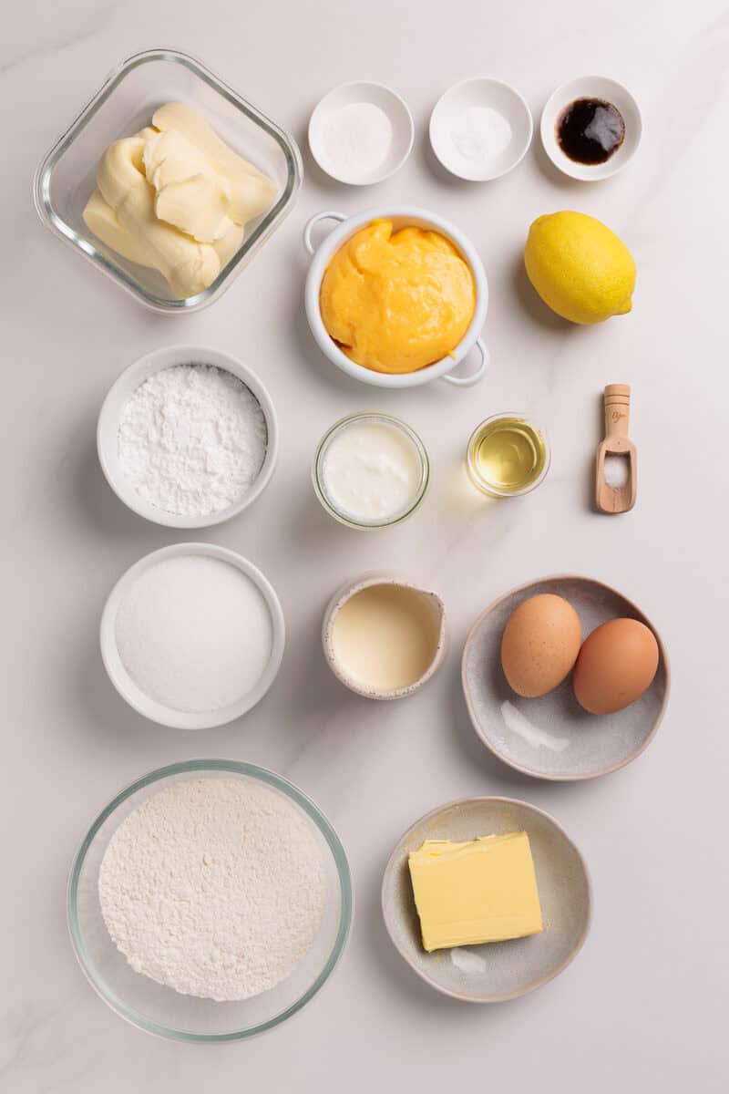 An overhead view of baking ingredients arranged neatly on a white surface, including flour, sugar, eggs, butter, lemon, custard, vanilla, powdered sugar, baking powder, salt, oil, and yogurt in various bowls and dishes.