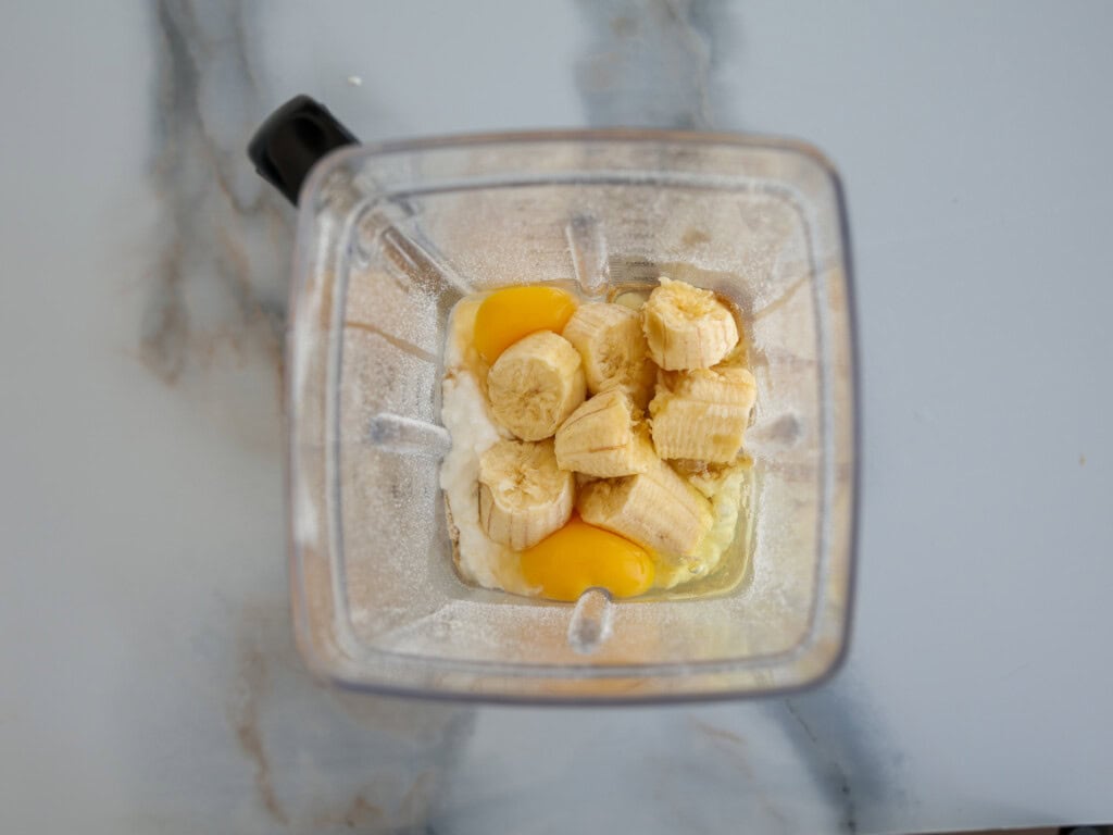 Overhead view of a blender containing chunks of banana, two cracked eggs, and a white creamy ingredient on a light countertop.