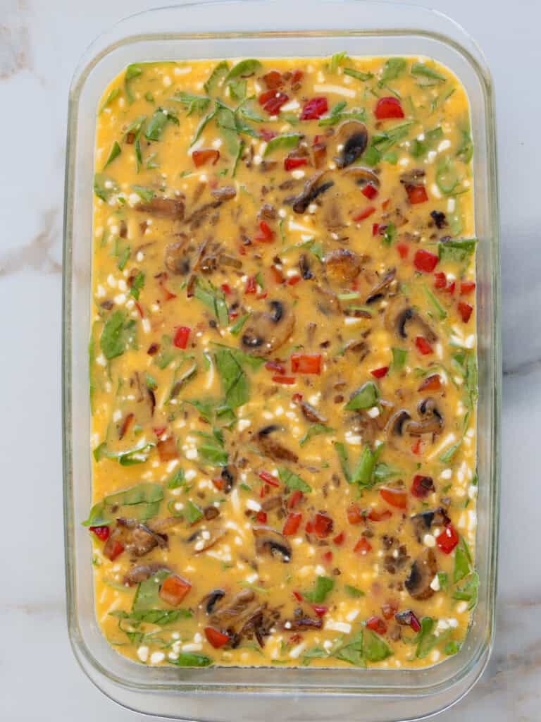 A glass baking dish filled with an uncooked egg mixture containing chopped spinach, bell peppers, mushrooms, and cheese, ready to be baked.