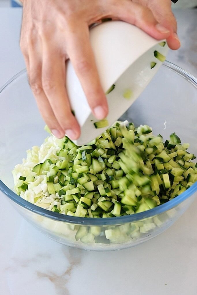 A hand pours chopped cucumber from a white bowl into a glass mixing bowl already containing chopped vegetables, likely cabbage, on a light-colored countertop.