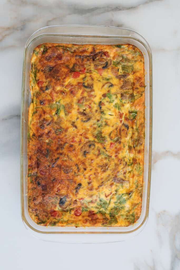 A glass baking dish filled with a golden-brown baked egg casserole containing visible pieces of vegetables, such as spinach, mushrooms, and red peppers, sits on a marble countertop.