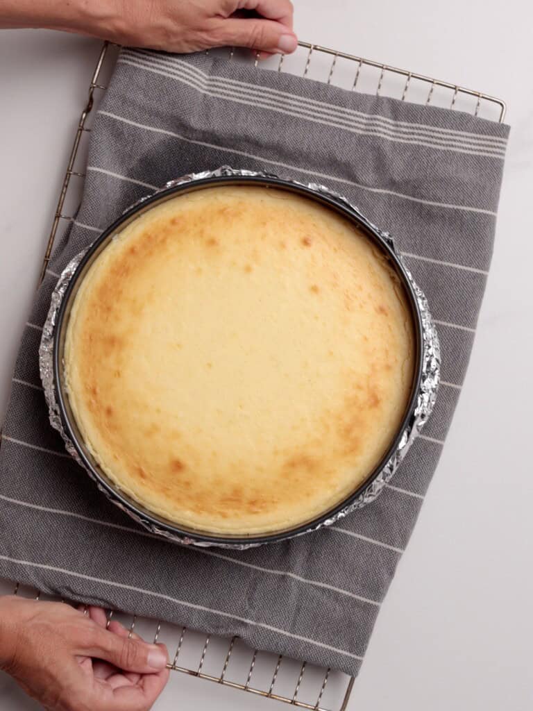 A round, baked cheesecake in a springform pan wrapped with foil sits on a gray striped kitchen towel atop a cooling rack. Two hands hold the rack at opposite corners.