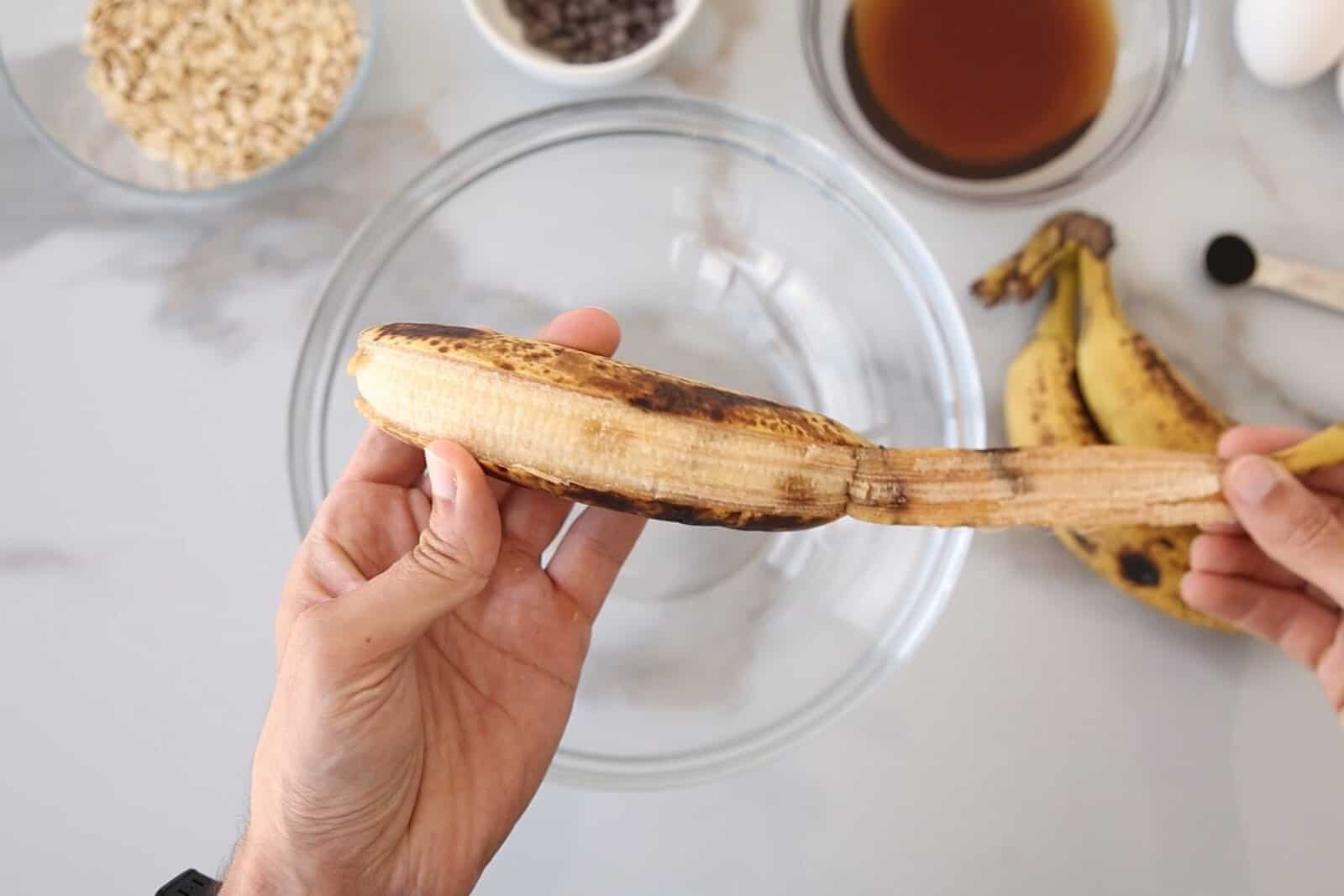 A person holds a partially peeled ripe banana over a glass mixing bowl, with ingredients like oats, chocolate chips, an egg, bananas, and vanilla extract in bowls nearby on a countertop.