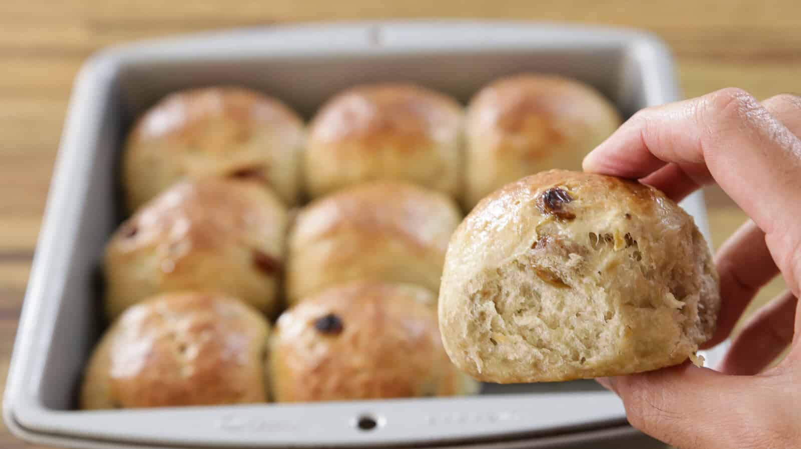 A hand holds a freshly baked banana bun above a baking pan filled with more golden brown buns, all with a shiny, glazed finish.