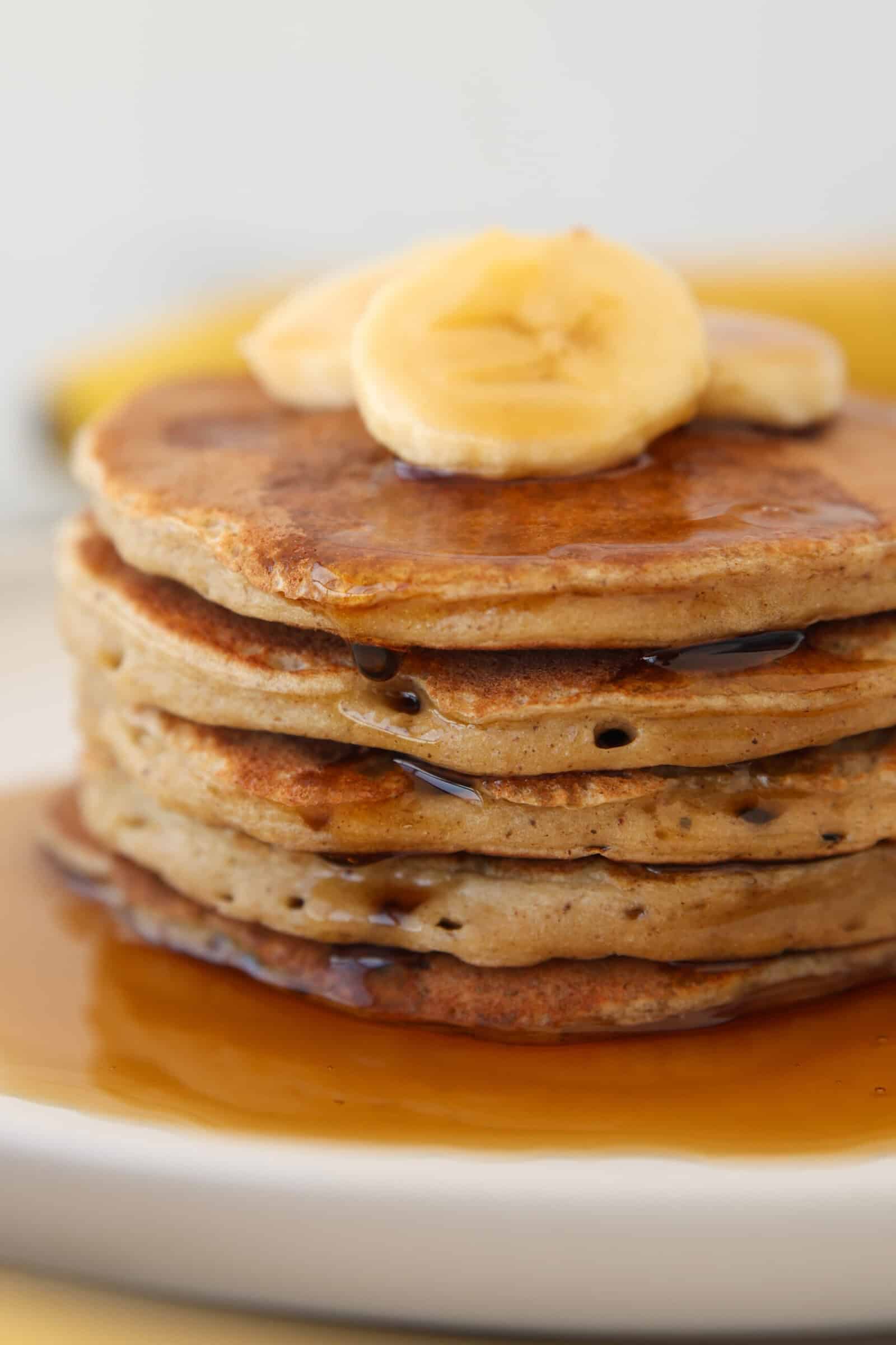 A stack of healthy banana and cottage cheese pancakes topped with banana slices and drizzled with syrup, served on a white plate.