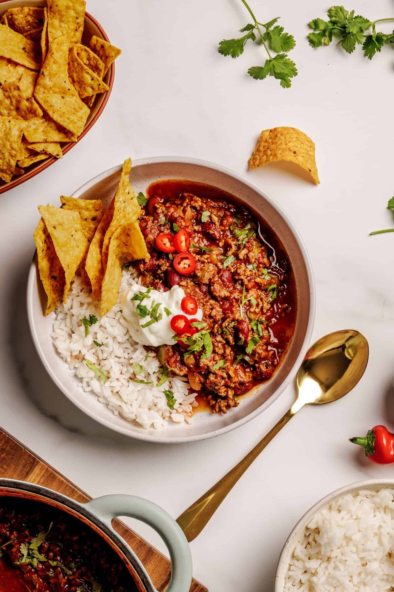 A bowl of chili con carne served with white rice, tortilla chips, sliced red chili peppers, fresh cilantro, and a dollop of sour cream, with a gold spoon on the side. Extra chips and rice are nearby on a white surface.