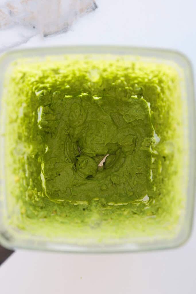 A top-down view of a blender containing a thick, smooth green mixture, likely a puree or sauce, with some of the mixture clinging to the sides.