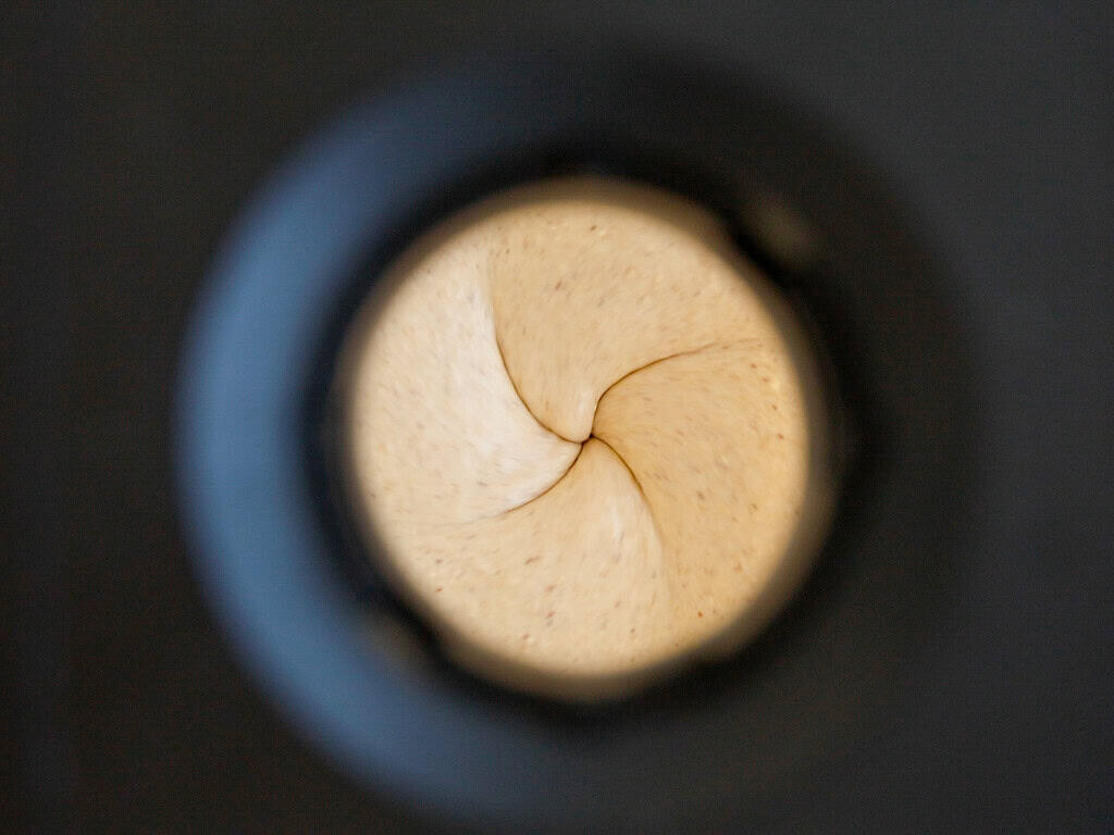 A top-down view through a blender lid, showing beige, swirled batter or dough being mixed inside, with the edges of the black blender opening framing the circular motion.
