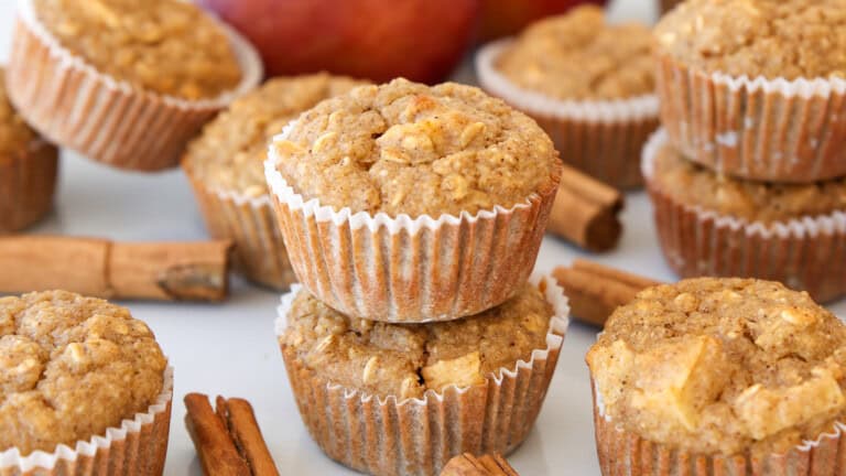 Close-up of apple breakfast oatmeal muffins stacked on a white surface, surrounded by cinnamon sticks, with whole apples blurred in the background. The muffins are golden brown and in white paper liners.