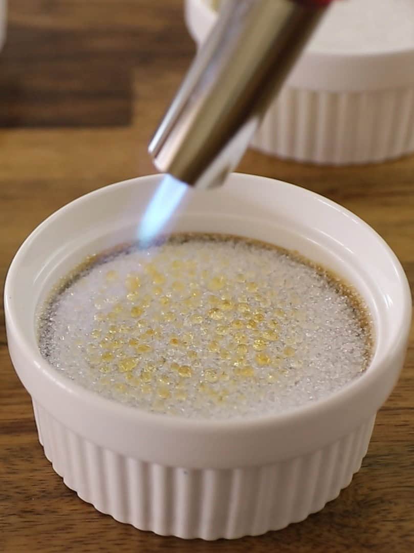 A kitchen torch caramelizes sugar on top of a pistachio-infused crème brûlée in a white ramekin, creating a golden, crisp layer. The dessert sits on a wooden surface.