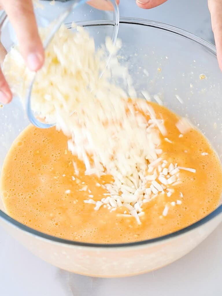 A hand is pouring shredded cheese from a small bowl into a large glass bowl filled with beaten eggs.