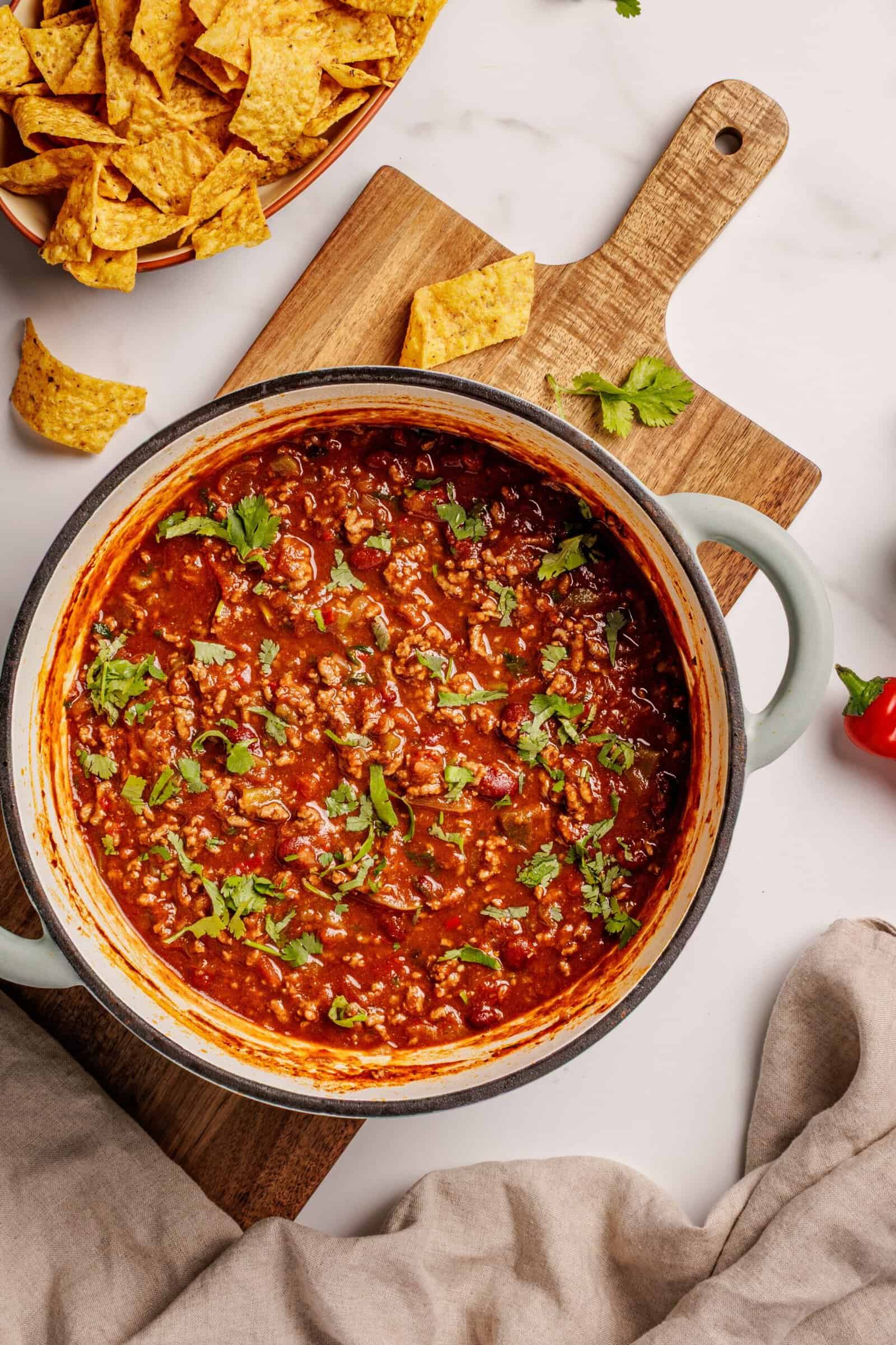 A pot of chili topped with chopped cilantro sits on a wooden board, surrounded by tortilla chips and a red chili pepper on a marble surface.