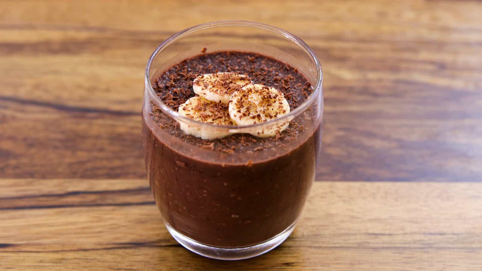 A glass filled with chocolate banana chia pudding, topped with banana slices and a sprinkle of chocolate shavings, sits on a wooden surface.