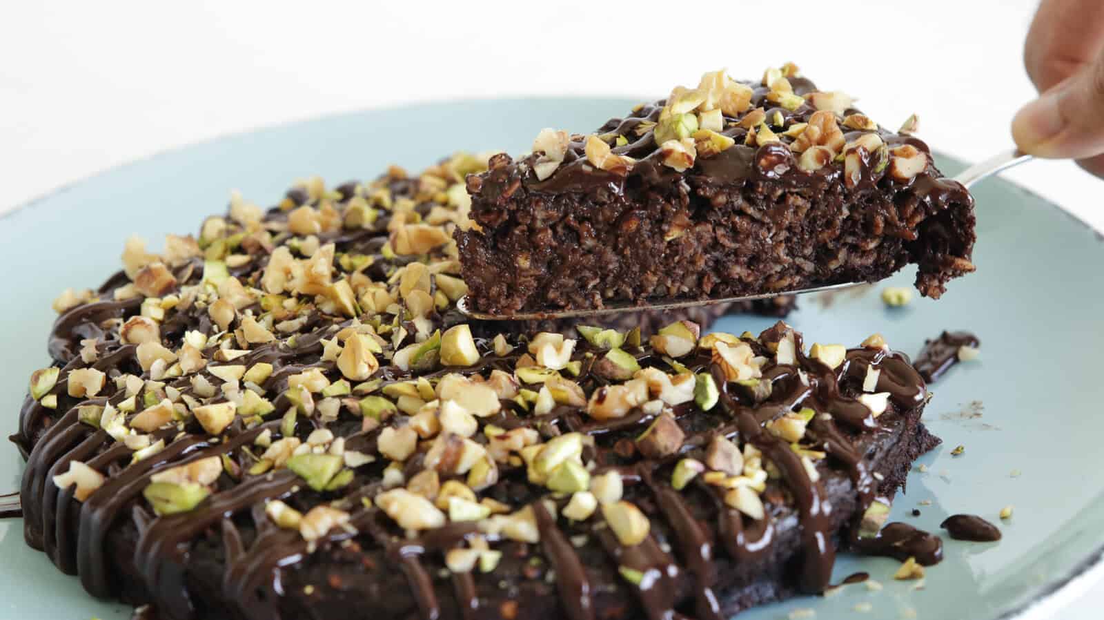 A close-up of a chocolate banana and oatmeal cake topped with chopped nuts and chocolate drizzle, with one slice being lifted from the cake on a spatula. The cake sits on a light blue plate.