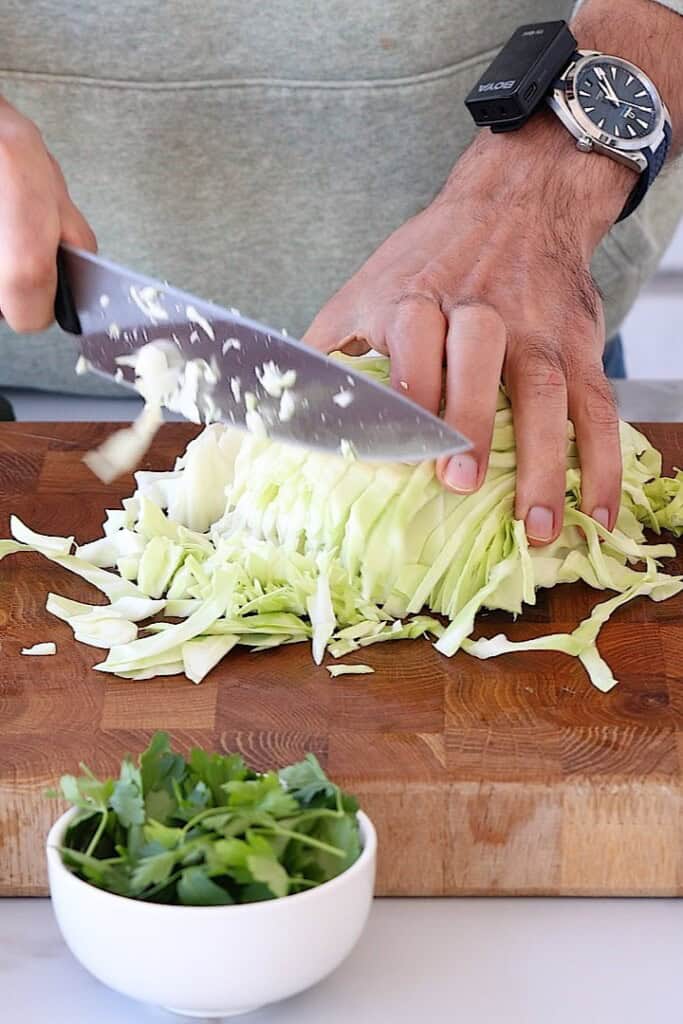 A person slices a head of cabbage on a wooden cutting board with a large knife. A small bowl of fresh green herbs sits in the foreground. The person wears a gray shirt and a watch.