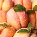 Sugared Italian peach-shaped cookies with a pink and orange hue, decorated with green mint leaves, closely arranged together.