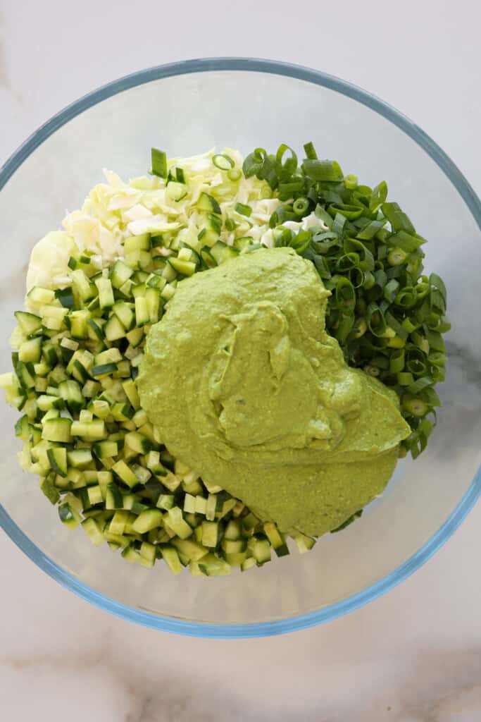 A glass bowl containing chopped green onions, cubed cucumber, shredded cabbage, and a large dollop of creamy green dressing, all ready to be mixed together.