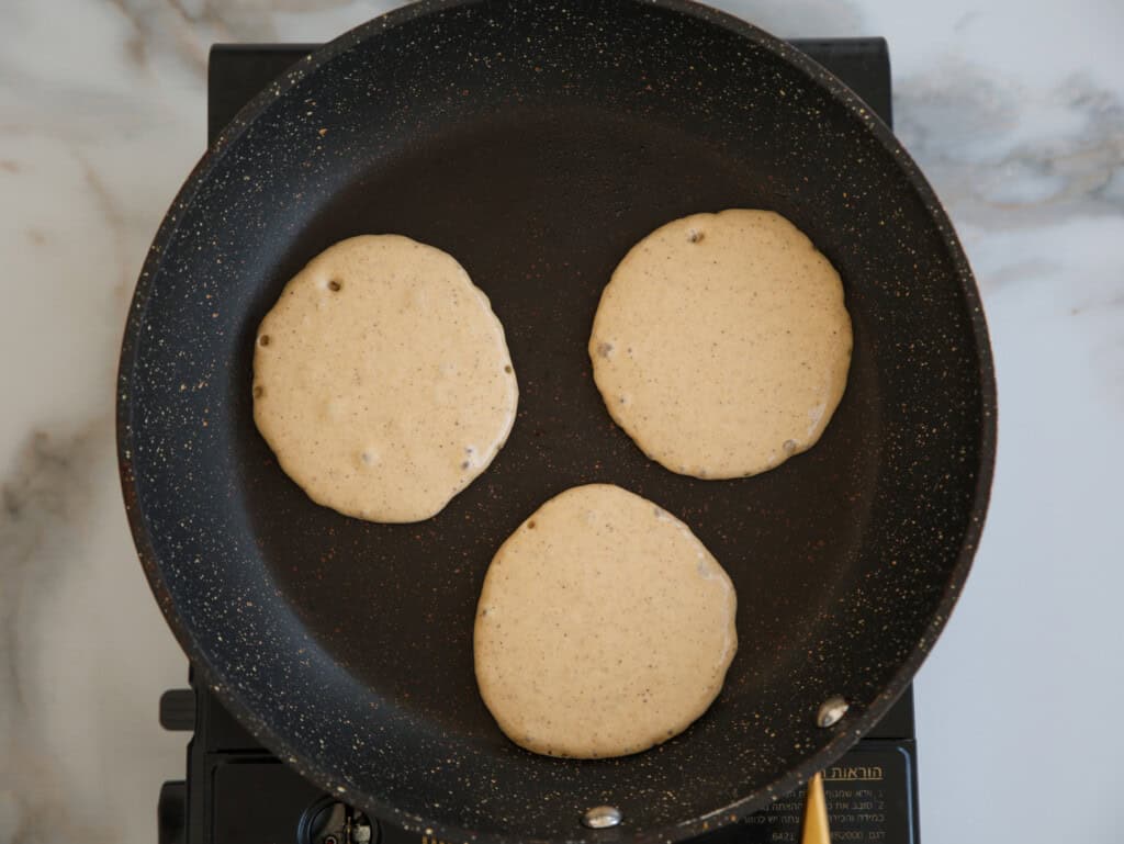 Three pancake batter rounds are cooking in a nonstick frying pan on a stove, viewed from above against a light marble background.